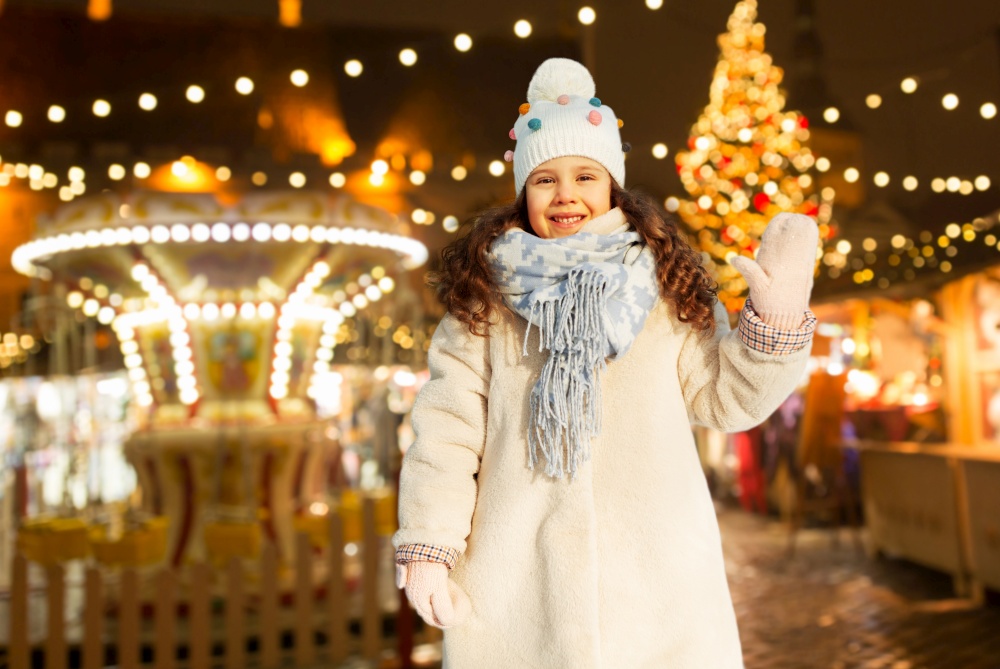childhood, leisure and winter holidays concept - portrait of happy little girl waving hand over night christmas market background. happy little girl waving hand at christmas market
