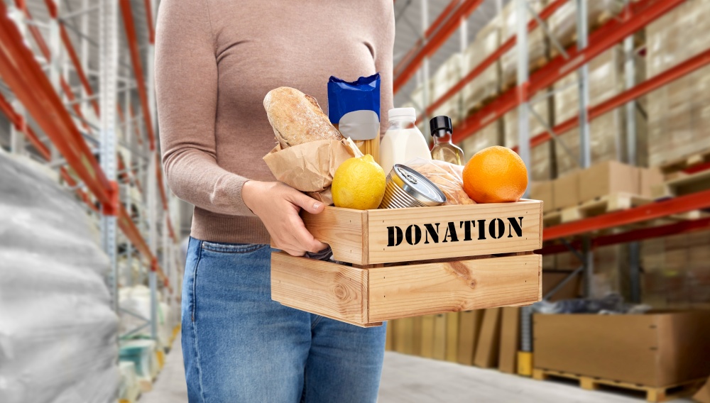 charity, relief work and people concept - close up of woman with food donation in wooden box over warehouse background. woman holds box with food donation at warehouse