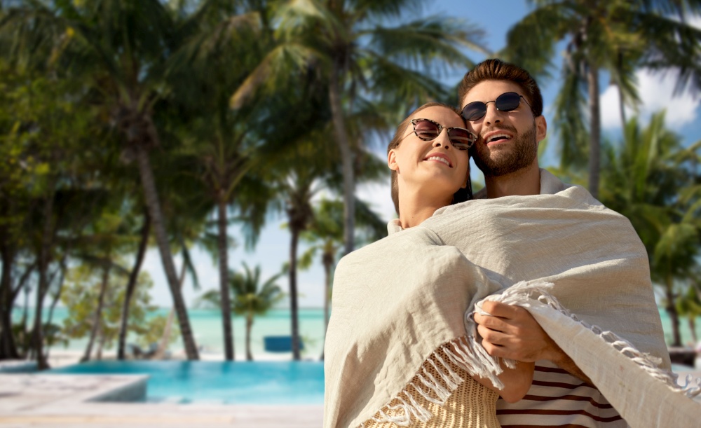 travel, tourism and people concept - happy couple in sunglasses covered with blanket hugging over tropical beach background in french polynesia. happy couple covered with blanket hugging on beach