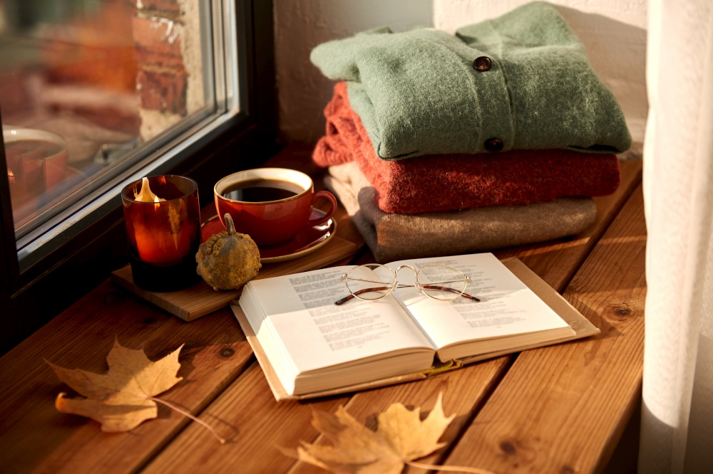 season and objects concept - open book with glasses, cup of coffee, autumn wool sweaters, pumpkin and candle burning on wooden window sill at home. book, coffee and autumn sweaters on window sill
