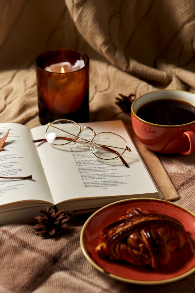 autumn, season and leisure concept - open book of poems with glasses, cup of coffee, cinnamon bun and candle on warm blankets at home. book, cinnamon bun, coffee and candle in autumn