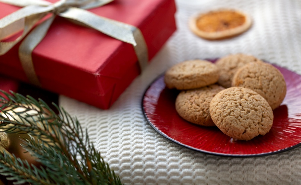 bake, food and winter holidays concept - close up of oatmeal cookies on red ceramic plate and christmas gift at home. cookies on plate and christmas gift at home
