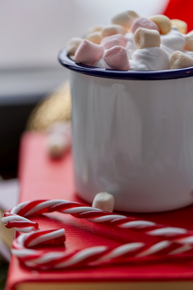 holidays, christmas and celebration concept - close up of camp mug with whipped cream and marshmallow, candy canes, books and decorations at home. cup of whipped cream with marshmallow on christmas