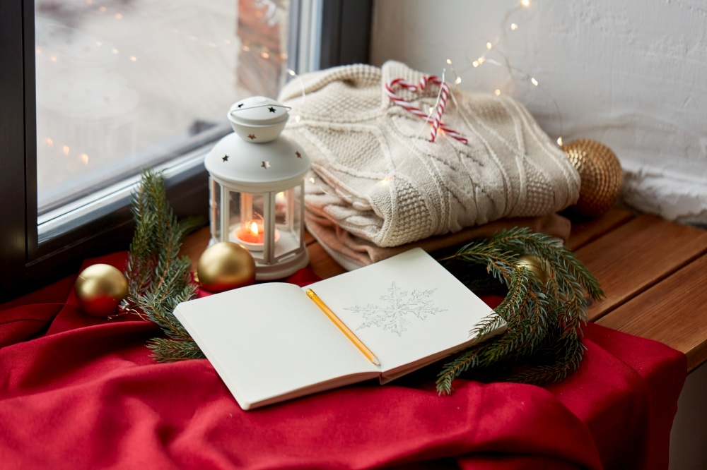 christmas, holidays and objects concept - close up of sketchbook with pencil drawing of snowflake, warm wool braided sweater, lantern and fir branch on red tablecloth on window sill at home. sketchbook, christmas lantern, sweater, fir branch
