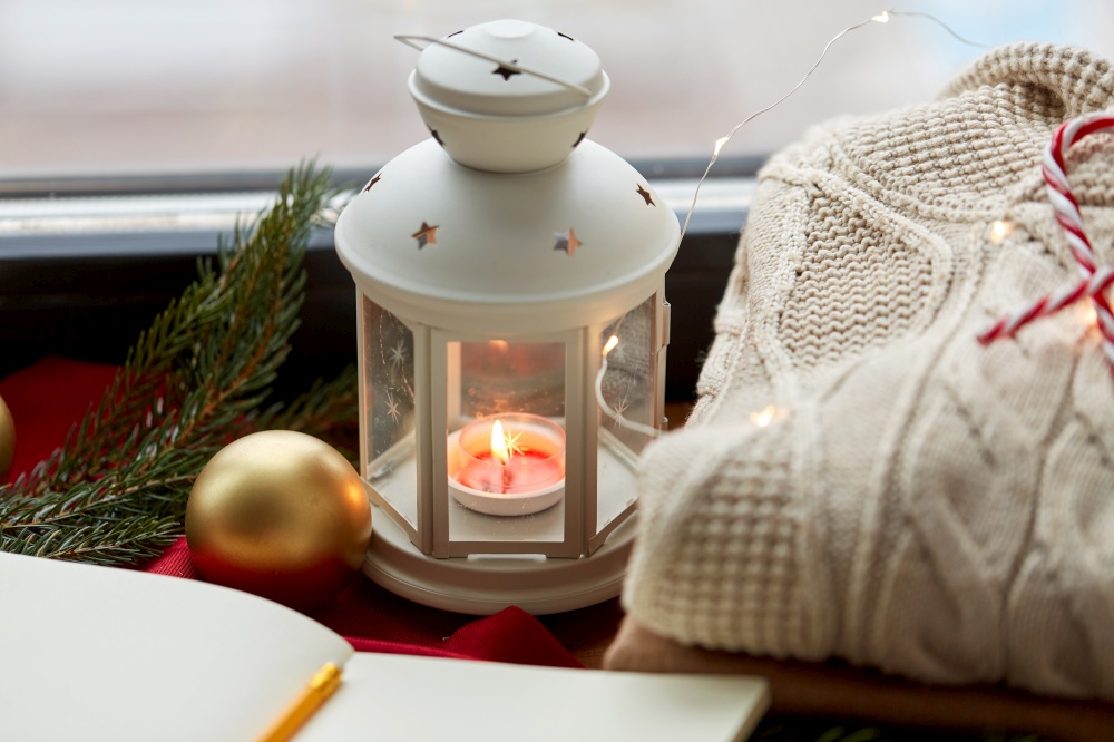 christmas, holidays and objects concept - close up of lantern with burning candle, warm wool braided sweater and sketchbook on window sill at home. candle burning in christmas lantern and sweater