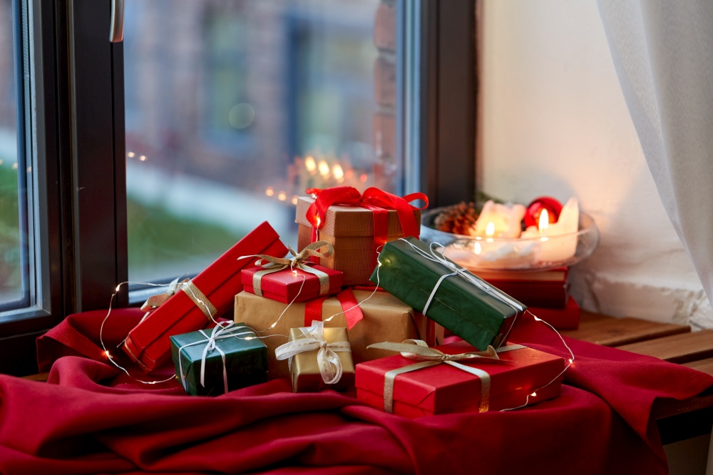 holidays, new year and celebration concept - heap of christmas gifts with electric garland on red tablecloth on window sill. christmas gifts on red tablecloth on window sill
