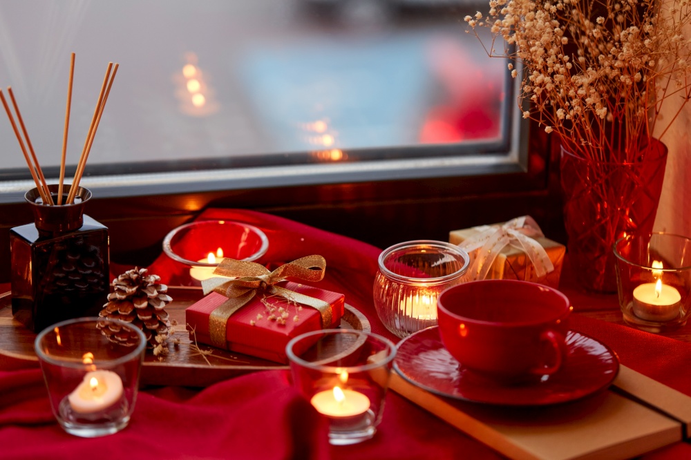 holidays, decoration and celebration concept - christmas gift, coffee cup cup, candles and aroma reed diffuser on red tablecloth on window sill at home. christmas gift, candles and cup on window sill