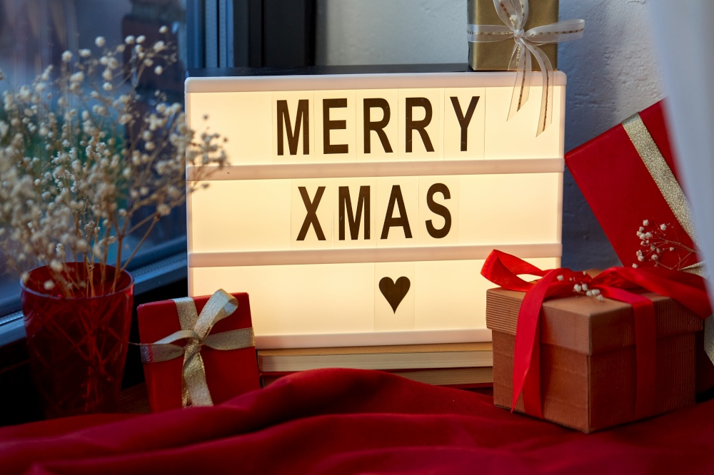holidays, decoration and celebration concept - merry christmas greeting on light box and gift boxes on red tablecloth on window sill at home. merry christmas on light box and gifts on window