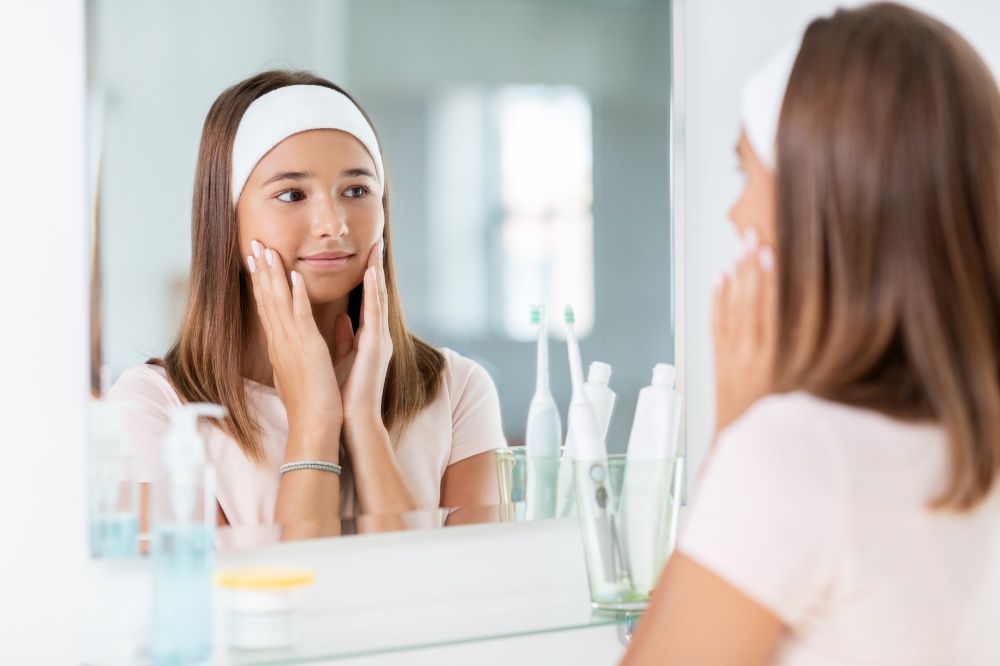 beauty, hygiene and people concept - teenage girl touching her face and looking in mirror at bathroom. teenage girl looking in mirror at bathroom