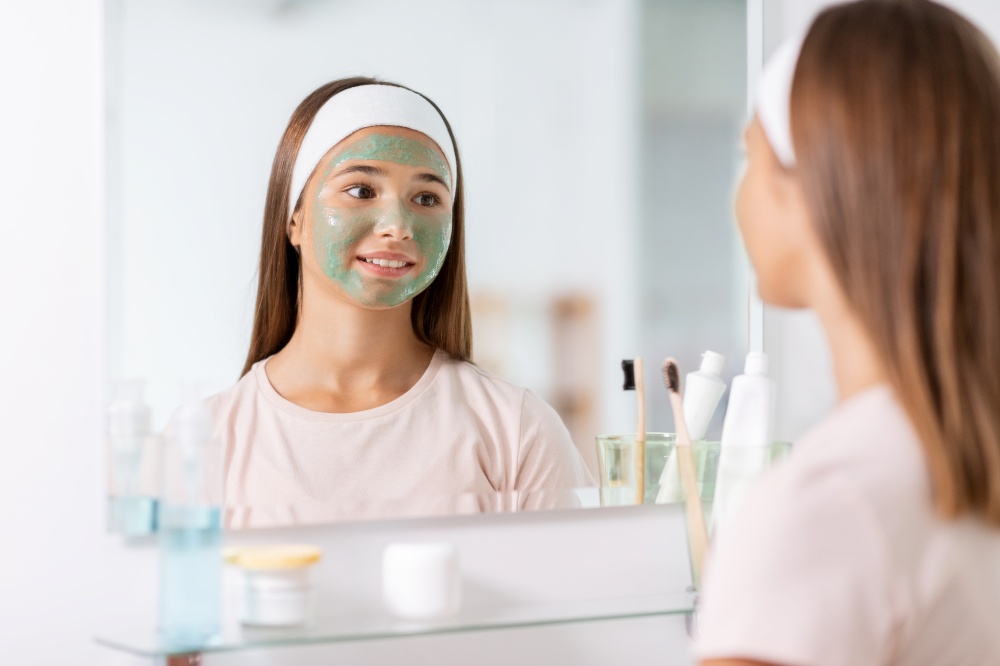 beauty, skin care and people concept - teenage girl with clay mask on her face looking in mirror at bathroom. girl with clay mask on face looking in mirror