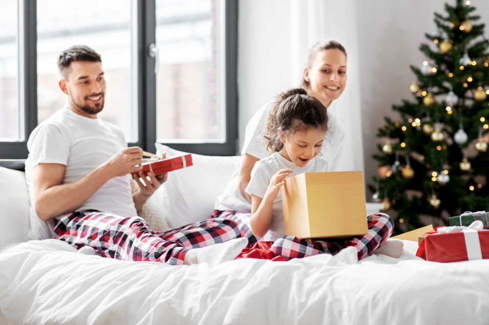 family, winter holidays and people concept - happy mother, father and little daughter opening christmas gifts in bed at home. happy family with christmas gifts in bed at home