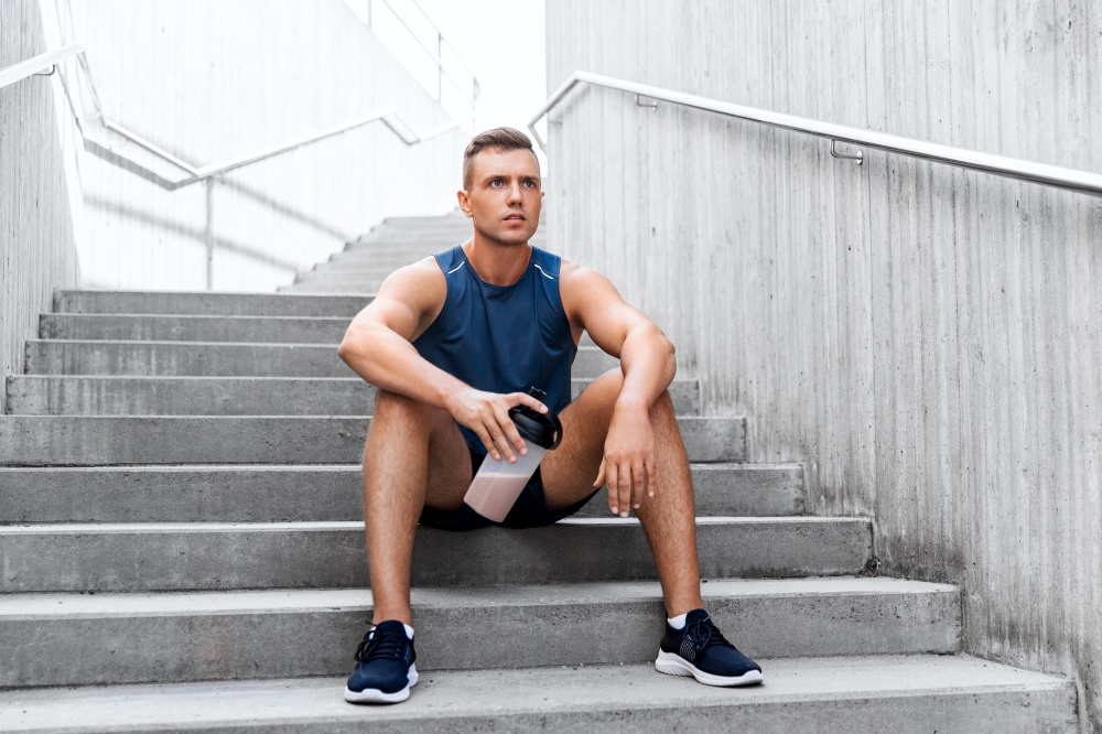 fitness, sport and people concept - tired young man with protein shake in bottle sitting on stairs. sportsman with shake in bottle sitting on stairs