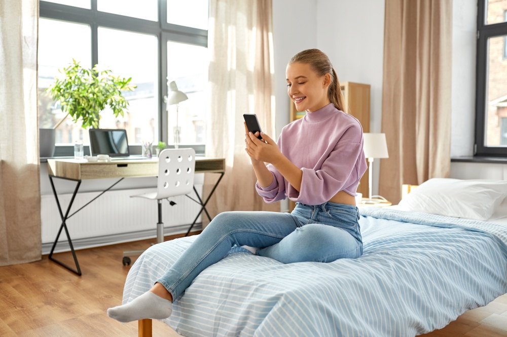 children, technology and communication concept - smiling teenage girl with smartphone sitting on bed at home. happy girl with smartphone sitting on bed at home