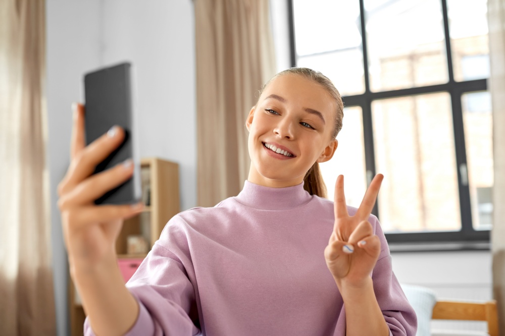 people, children and technology concept - happy smiling teenage girl with smartphone taking selfie and showing peace gesture at home. happy girl with smartphone taking selfie at home