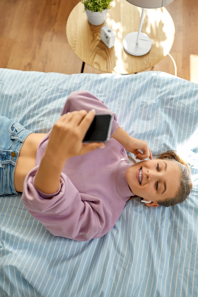 leisure, children and technology concept - happy smiling girl in earphones with smartphone lying on bed at home. girl in earphones with smartphone lying on bed