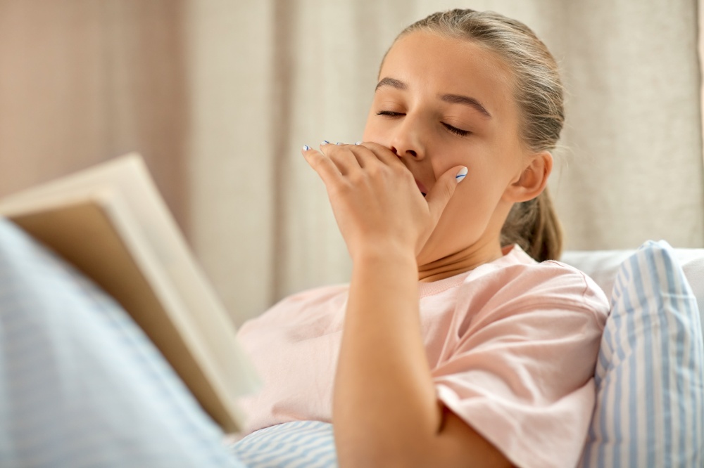 people, bedtime and sleeping concept - yawning girl reading book in bed at home. yawning girl reading book in bed at home