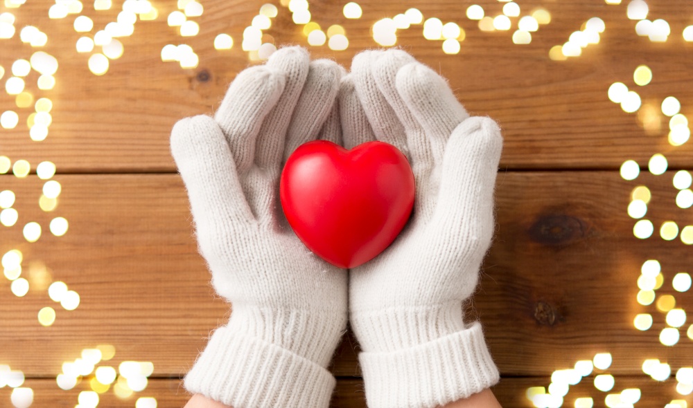 winter, valentine&rsquo;s day and christmas concept - hands in white woollen gloves holding red heart over wooden boards background with bokeh lights. hands in white woollen gloves holding red heart