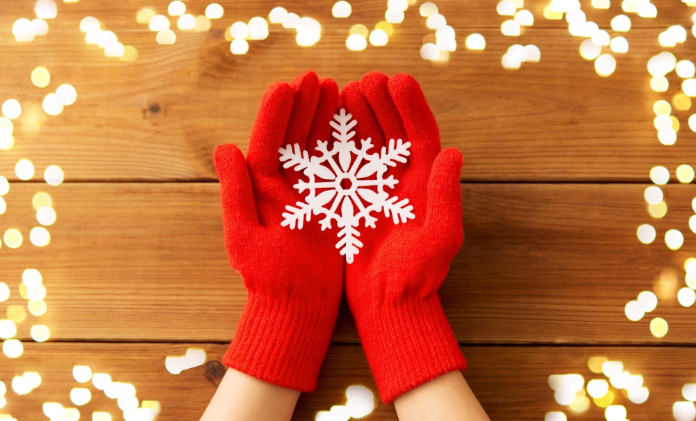 winter and christmas concept - hands in red woollen gloves holding big white snowflake over wooden boards background with bokeh lights. hands in red woollen gloves holding big snowflake
