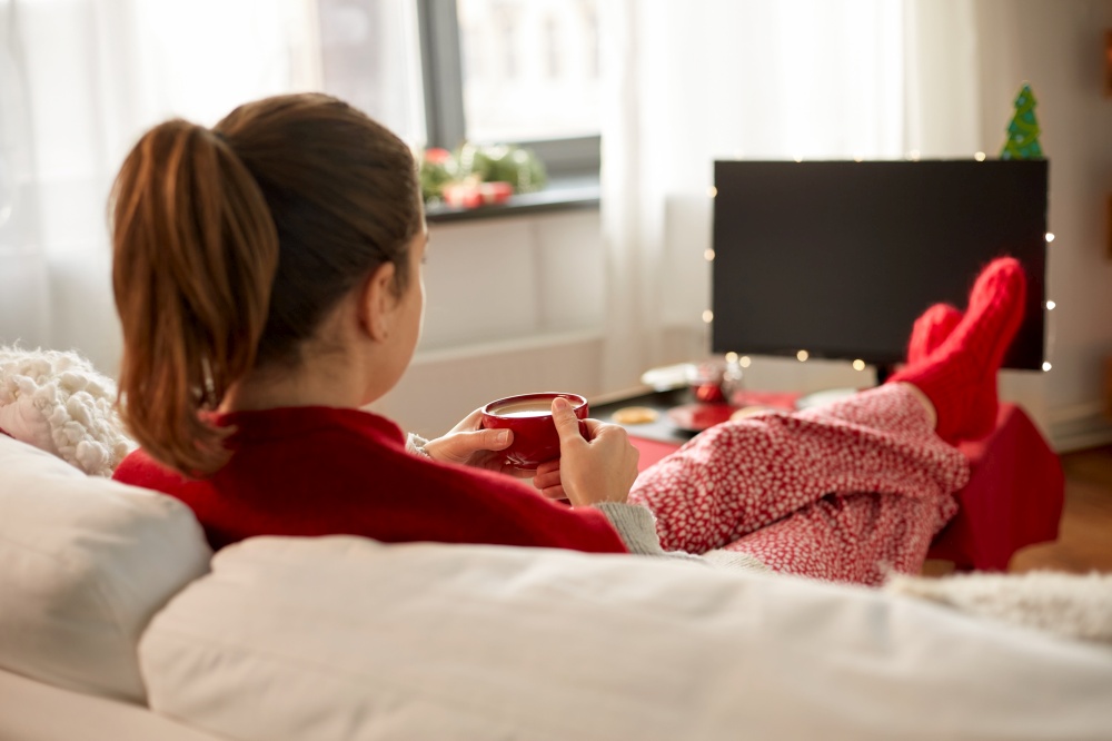 christmas, winter holidays and leisure concept - close up of young woman watching tv and drinking coffee with her feet on table at cozy home. woman watching tv and drinking coffee on christmas