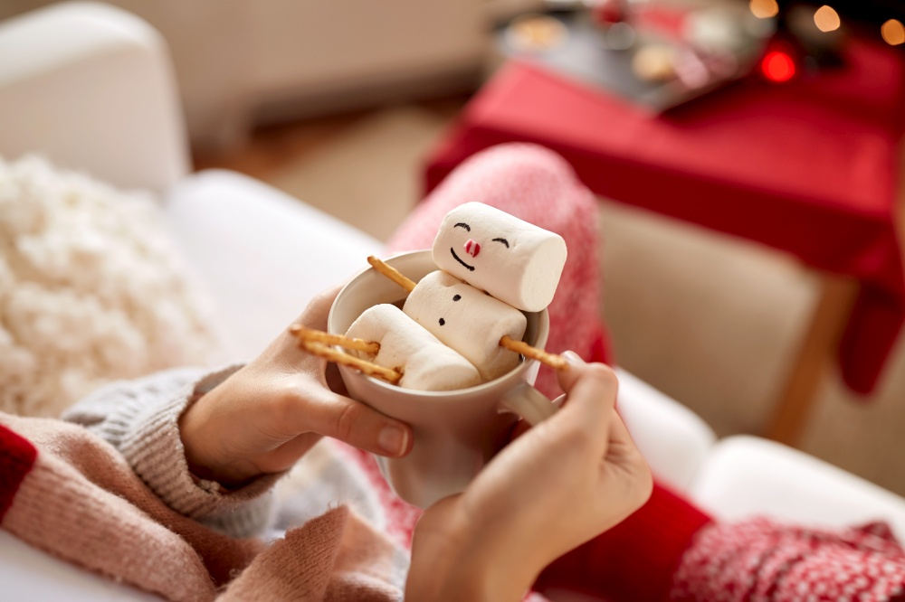 christmas, winter holidays and leisure concept - close up of hands holding mug with marshmallow snowman at home. hands with marshmallow snowman in mug on christmas