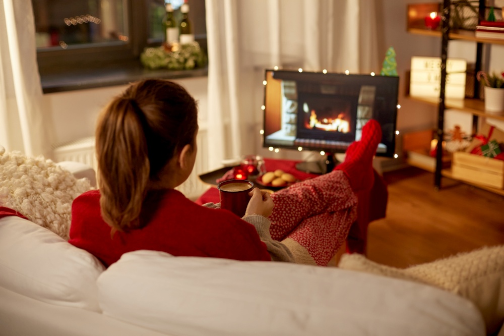 christmas, winter holidays and leisure concept - young woman watching tv with fireplace on screen and drinking coffee with her feet on table at cozy home. woman watching tv and drinking coffee on christmas
