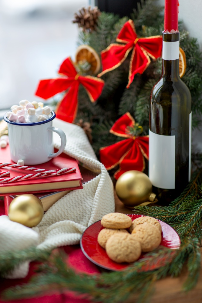 winter holidays concept - cup of whipped cream with marshmallow and candy canes, books, oatmeal cookies, candle in wine bottle and christmas decorations on window sill at home. christmas treats and decorations on window sill