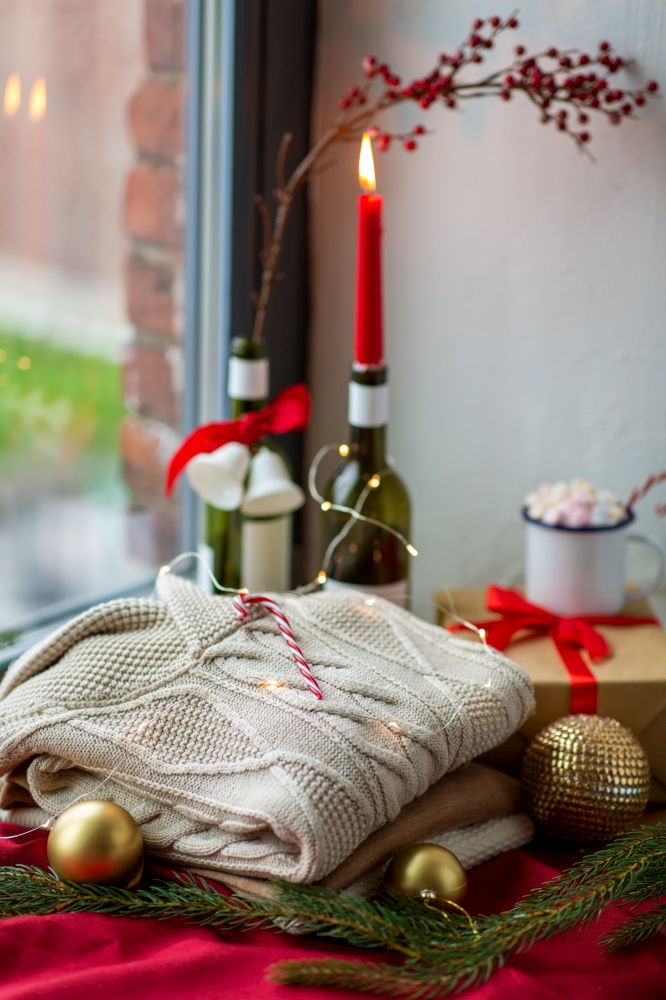 winter holidays concept - close up of warm wool braided sweater, candy cane, electric garland string, christmas decorations and candle burning in wine bottle on window sill at home. close up of sweater and christmas decor on sill