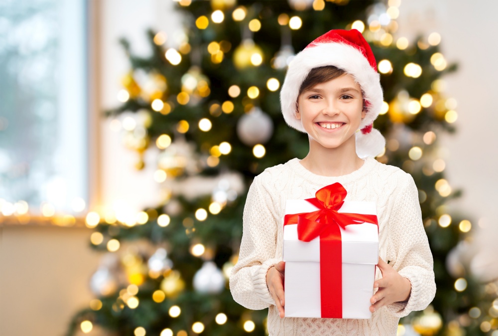 winter holidays, celebration and people concept - happy smiling boy in santa hat with gift box over christmas tree lights background. smiling boy in santa hat with christmas gift
