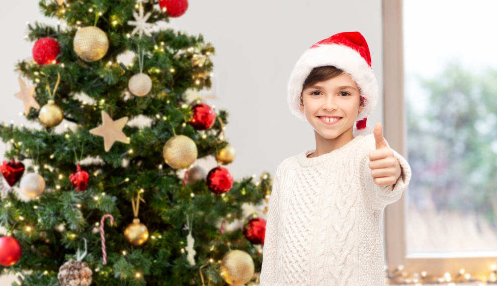 winter holidays, gesture and people concept - smiling happy boy in santa hat showing thumbs up over christmas tree on background. happy boy showing thumbs up over christmas tree