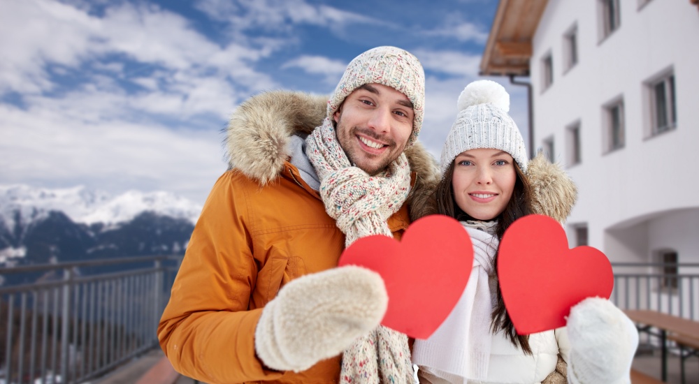 people, love and valentine&rsquo;s day concept - happy couple holding blank red hearts over winter resort on background. happy couple with red hearts over winter resort