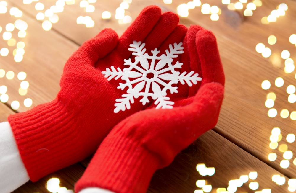 winter and christmas concept - hands in red woollen gloves holding big white snowflake over wooden boards background with bokeh lights. hands in red woollen gloves holding big snowflake
