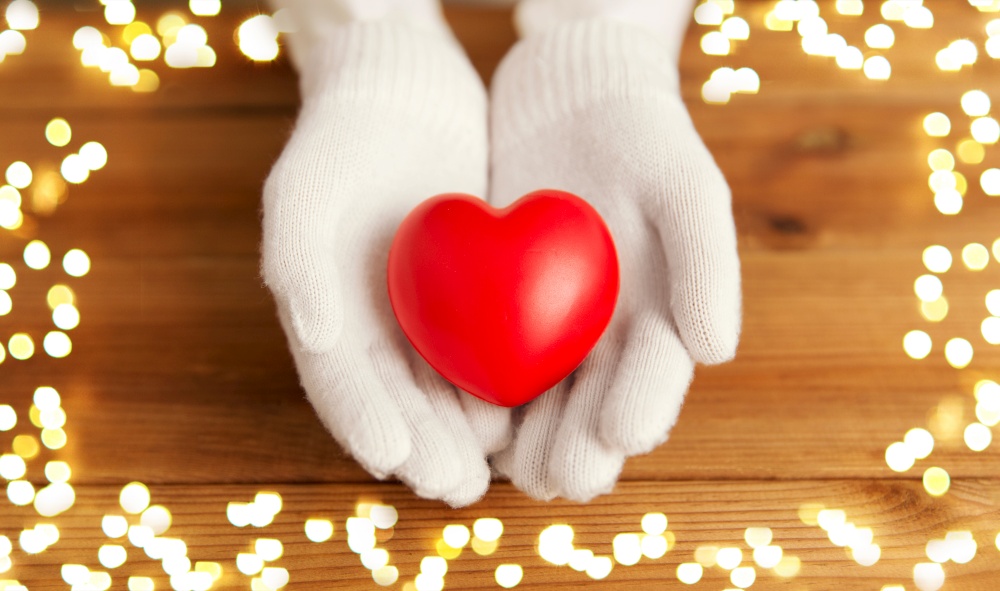 winter, valentine&rsquo;s day and christmas concept - hands in pale pink woollen gloves holding red heart over wooden boards background with bokeh lights. hands in pink woollen gloves holding red heart