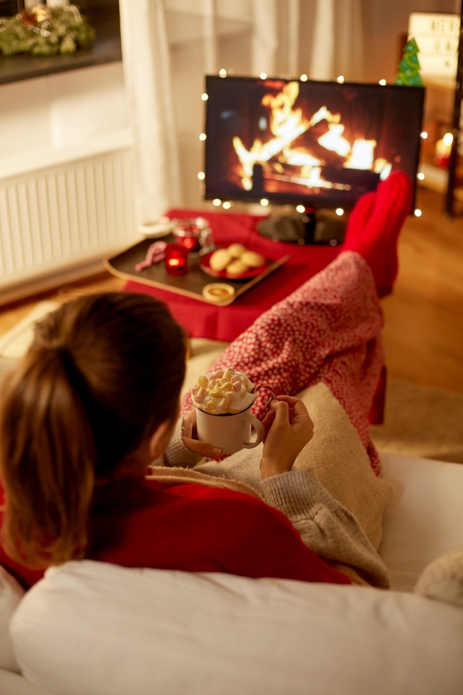 christmas, winter holidays and leisure concept - young woman watching tv with fireplace on screen and holding mug of marshmallow and whipped cream resting feet on table at cozy home. woman watching tv with marshmallow on christmas