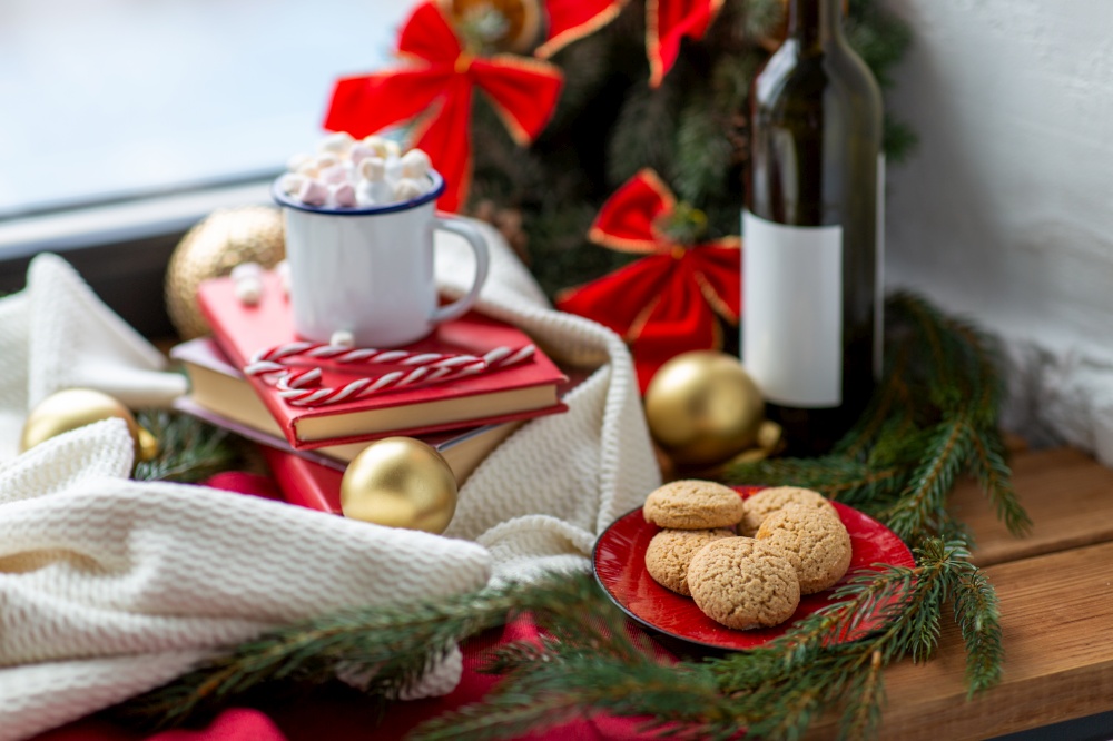 winter holidays concept - oatmeal cookies, cup of whipped cream with marshmallow and candy canes, books, wine bottle and christmas decorations on window sill at home. christmas treats and decorations on window sill