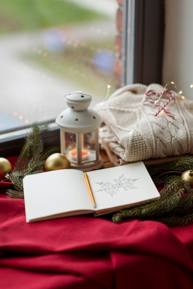 christmas, holidays and objects concept - close up of sketchbook with pencil drawing of snowflake, warm wool braided sweater, lantern and fir branch on red tablecloth on window sill at home. sketchbook, christmas lantern, sweater, fir branch