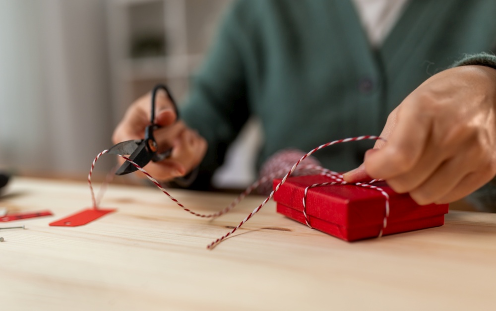 winter holidays and hobby concept - close up of woman with red box, rope and scissors packing christmas gift at home. close up of woman packing christmas gift at home