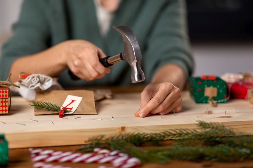 winter holidays and hobby concept - close up of woman with hammer hammering nails in wooden board and making advent calendar at home. woman making advent calendar on christmas at home