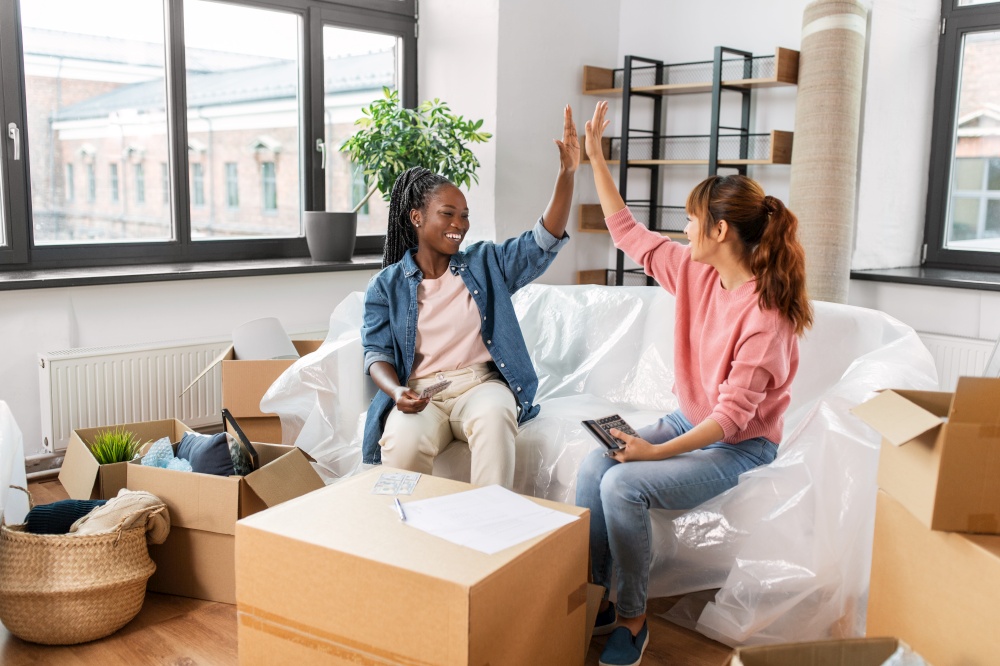 moving, finances and real estate concept - happy women with money, bills and calculator at new home making high five gesture. happy women moving to new home and counting money