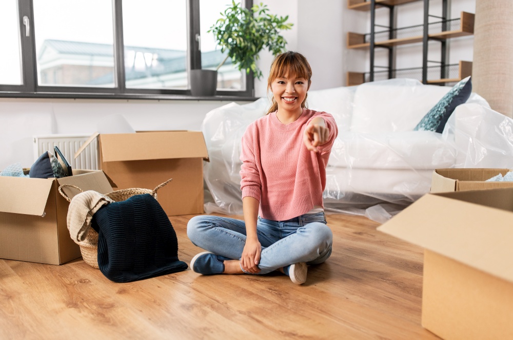moving, people and real estate concept - happy smiling woman with boxes sitting on floor at new home and pointing to camera. happy woman with boxes moving to new home