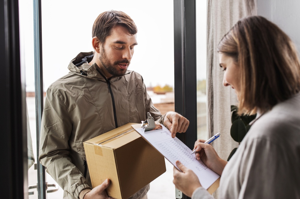 shipping, transportation and people concept - delivery man with parcel box and customer signing papers on clipboard at home. delivery man with parcel box and customer at home