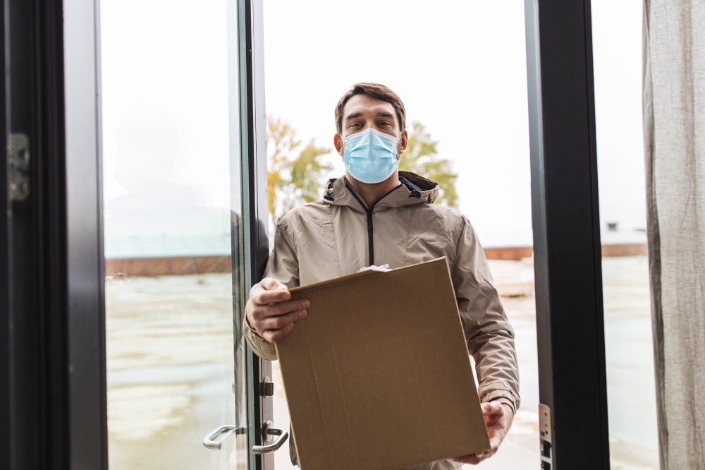 health protection, safety and pandemic concept - delivery man in face protective mask holding parcel box with clipboard at open door. delivery man in face mask holding parcel box