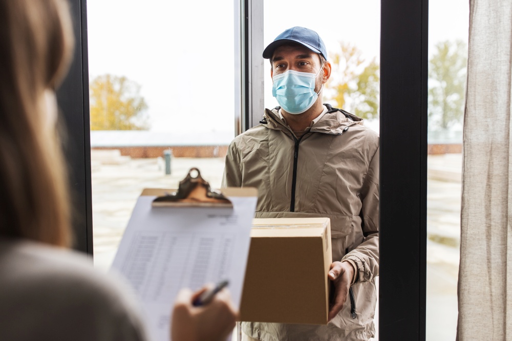 health protection, safety and pandemic concept - delivery man in face protective mask with parcel box and female customer signing papers at home. delivery man in mask with box and customer at home