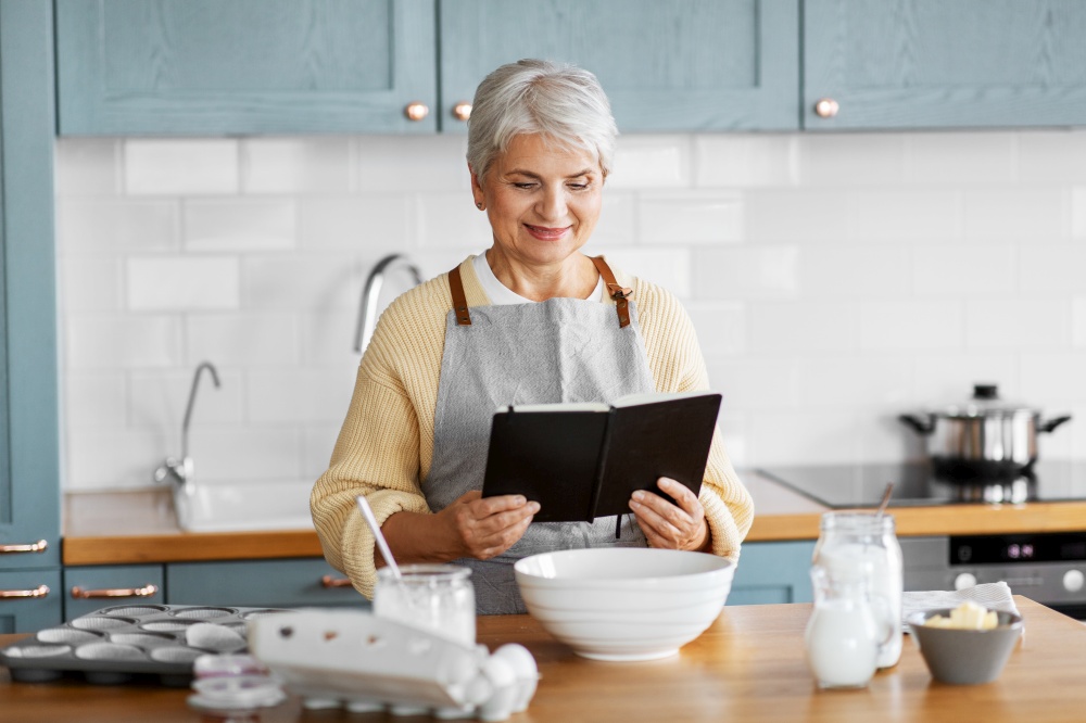 culinary, baking and people concept - happy smiling woman with cook book cooking food on kitchen at home. happy woman with cook book cooking food on kitchen