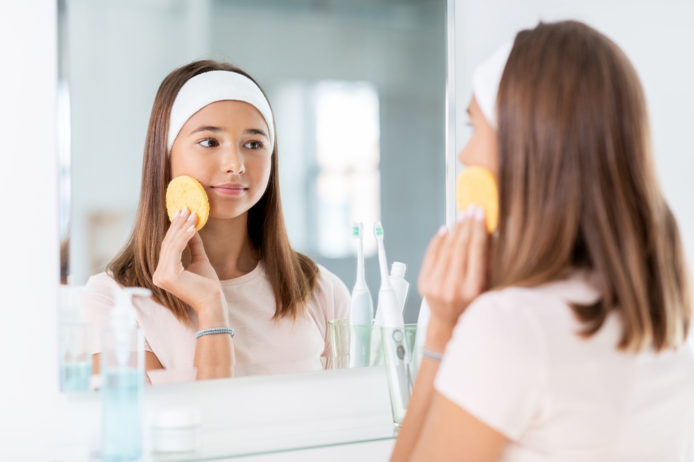 beauty, hygiene and people concept - teenage girl with cleansing sponge cleaning facial skin and looking in mirror at bathroom. teenage girl cleaning face with sponge at bathroom