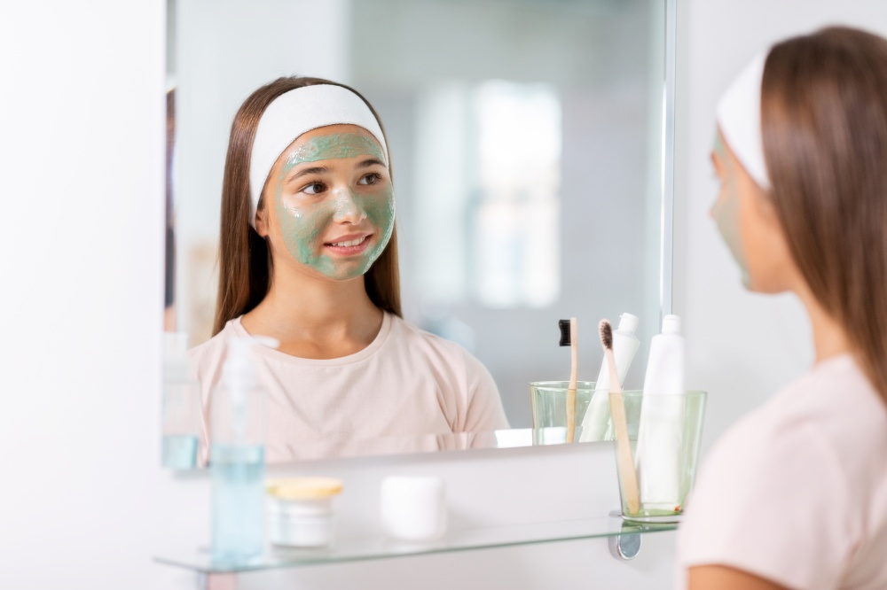 beauty, skin care and people concept - teenage girl with clay mask on her face looking in mirror at bathroom. girl with clay mask on face looking in mirror