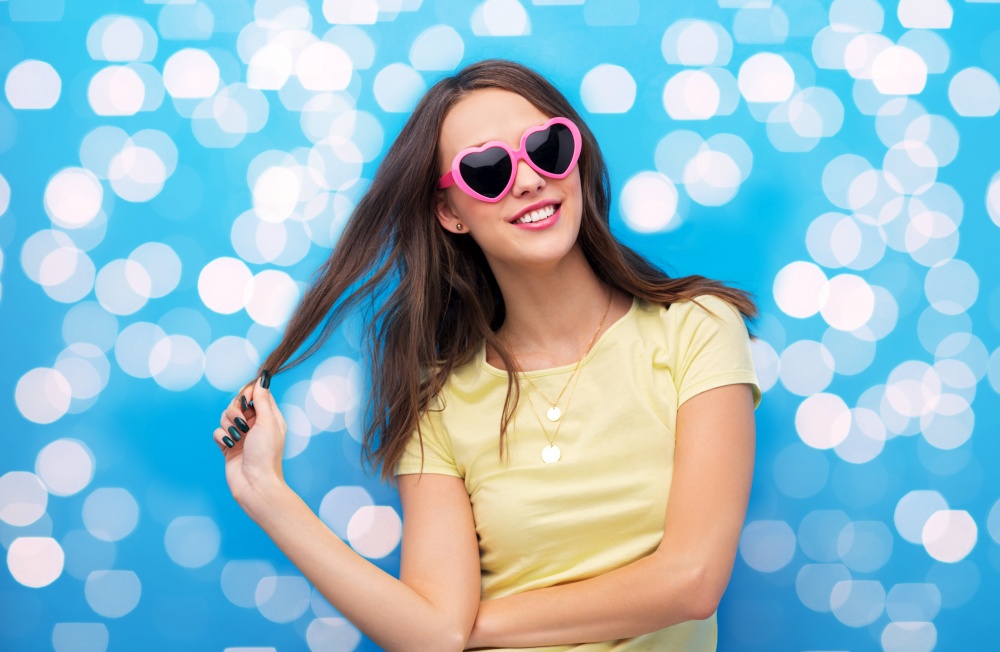 summer, valentine&rsquo;s day and people concept - smiling young woman or teenage girl in yellow t-shirt and heart-shaped sunglasses over lights on bright blue background. teenage girl in heart-shaped sunglasses on lights