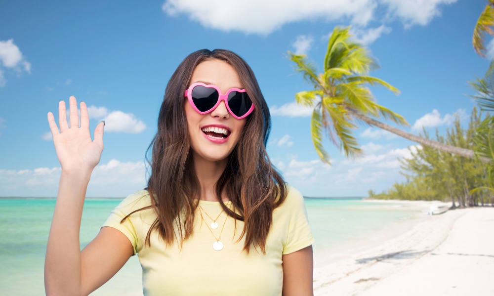 summer holidays, valentine&rsquo;s day and travel concept - smiling young woman or teenage girl in heart-shaped sunglasses waving hand over tropical beach background in french polynesia. happy woman in heart-shaped sunglasses on beach