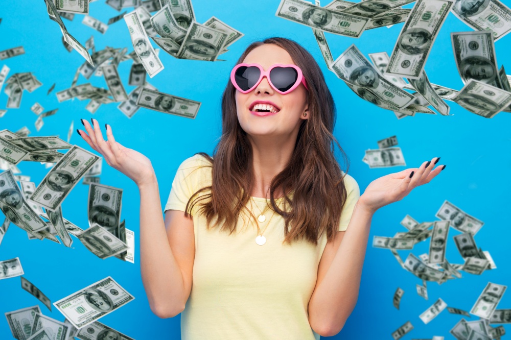 summer, valentine&rsquo;s day and people concept - smiling young woman or teenage girl in yellow t-shirt and heart-shaped sunglasses over dollar money rain on bright blue background. teenage girl in heart-shaped sunglasses over money