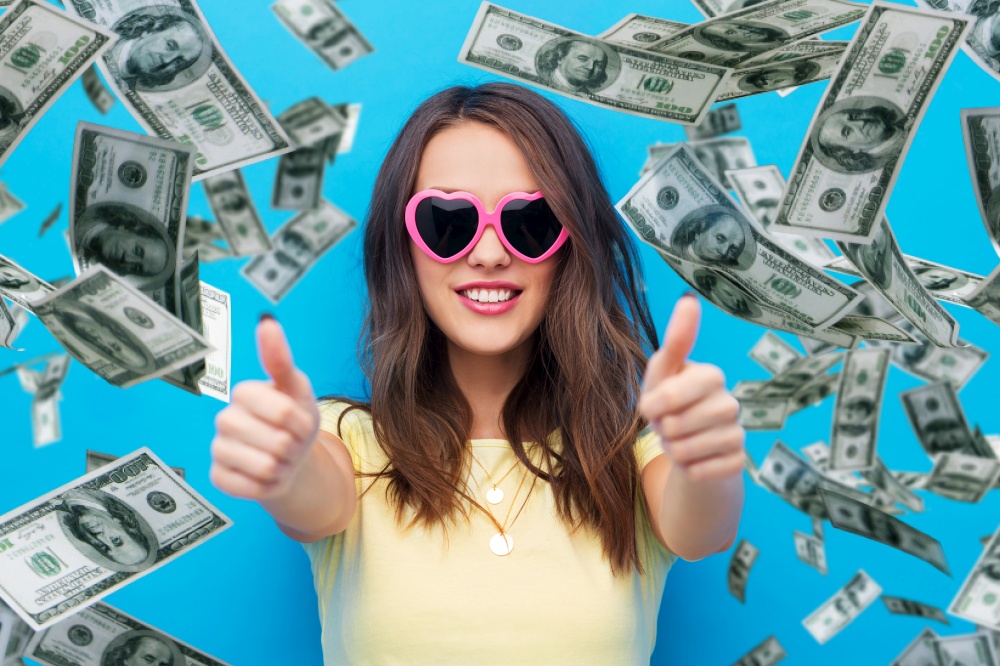 summer, valentine&rsquo;s day and people concept - smiling young woman or teenage girl in yellow t-shirt and heart-shaped sunglasses showing thumbs up over dollar money rain on bright blue background. woman in sunglasses shows thumbs up over money