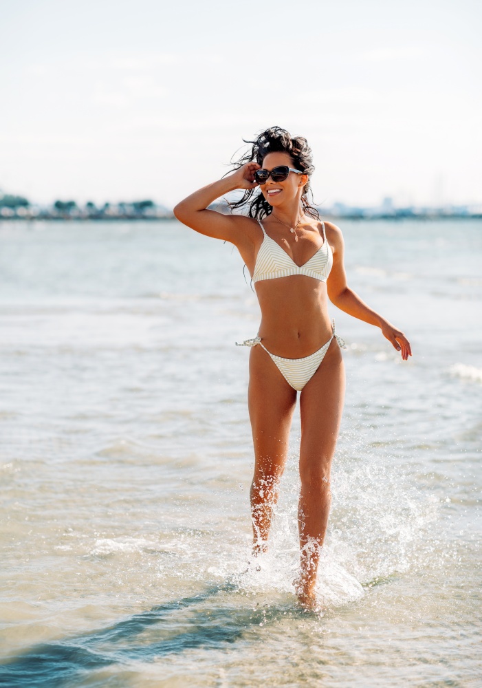 people, summer and swimwear concept - happy smiling young woman in bikini swimsuit running in water beach. smiling young woman in bikini swimsuit on beach